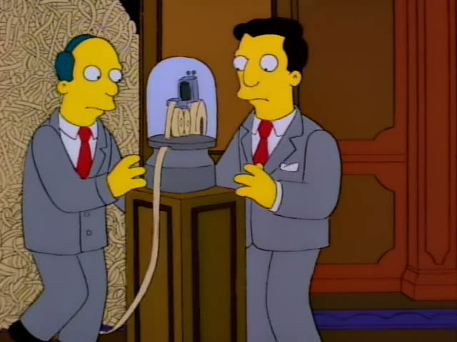 Two men in grey suits and red ties stand on either side of a glass dome on a wooden pedestal, with a cable running down…