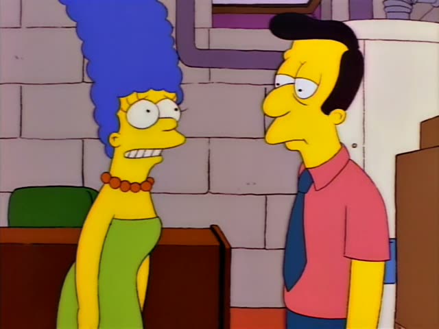 Marge Simpson stands at a wooden counter in a basement beside a brick wall and a white water heater, talking to an unnamed…
