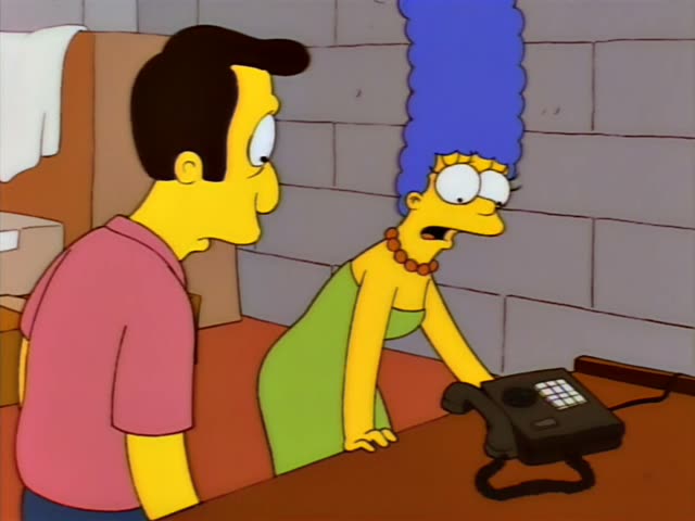 Marge Simpson stands at a counter beside a black telephone while a man in a pink shirt and dark tie, who resembles Ned-less…