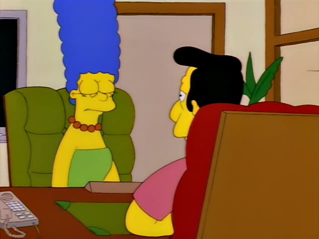 Marge Simpson stands beside a desk while a black-haired man in a pink shirt and blue tie sits in a red chair with his arms…