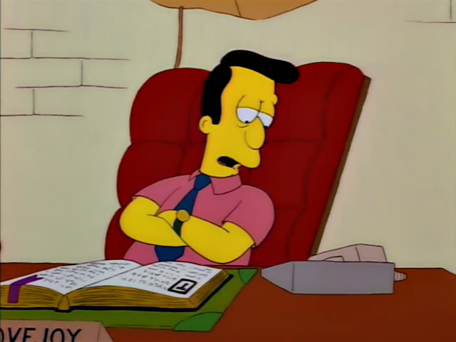 A black-haired man in a pink short-sleeved shirt and blue tie sits with his arms folded in a red armchair behind a wooden…