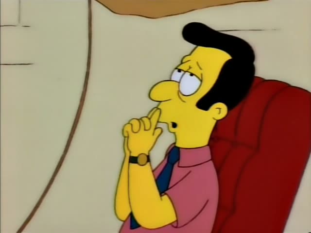 A slick-haired black-haired man in a pink shirt and blue tie sits in a red armchair against a plain wall, first…