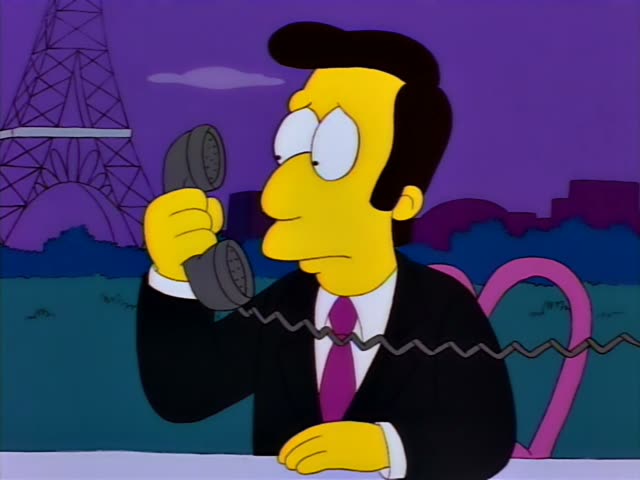 A slick-haired, yellow-skinned man in a black suit and magenta tie holds a black telephone receiver to his ear while sitting…
