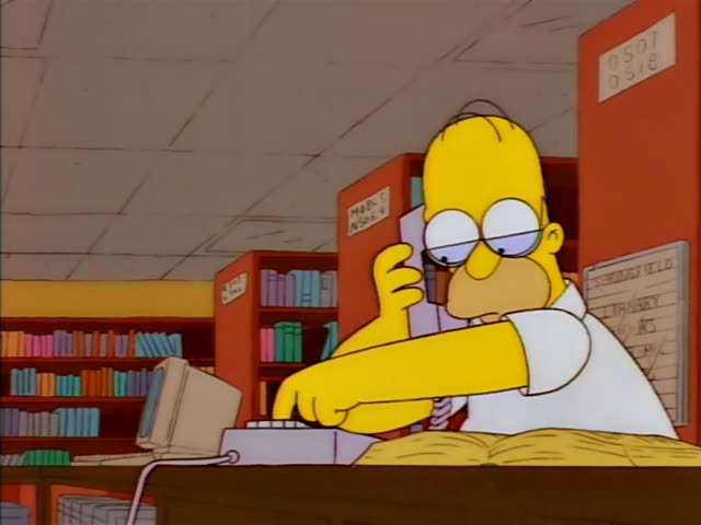 Homer Simpson sits at a desk in a library with shelves of colorful books behind him and a sign reading 0507 0.218 on the wall