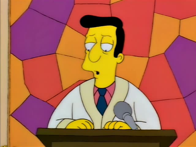A thin, dark-haired man in a white robe and blue tie stands behind a wooden podium with a microphone, speaking…