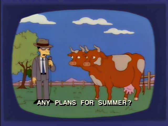 A man in a grey suit, brown fedora and glasses holds a microphone and a cord-linked device beside two brown-and-white cows…