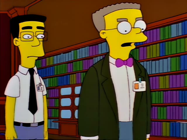Two men in a library lined with bookshelves stand together, a thin dark-haired man in a white shirt, black tie and glasses…