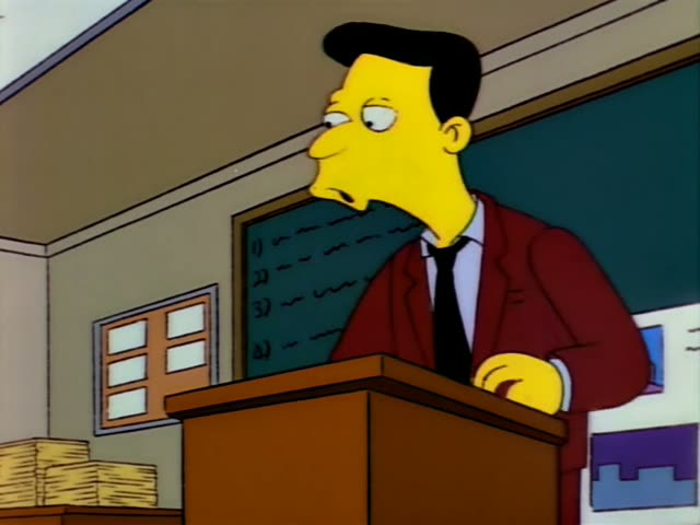 A balding-free man with black slicked-back hair, a red jacket and a black tie stands behind a wooden podium in a classroom…