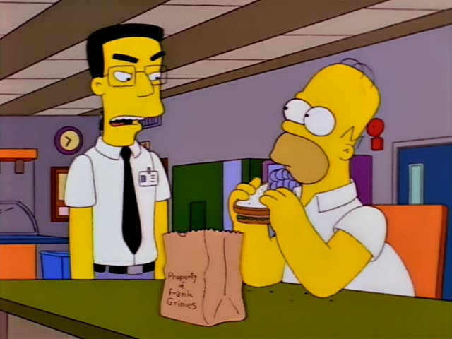 Homer Simpson stands at a counter eating a small sandwich while a thin man with black hair, glasses, a white shirt and black…