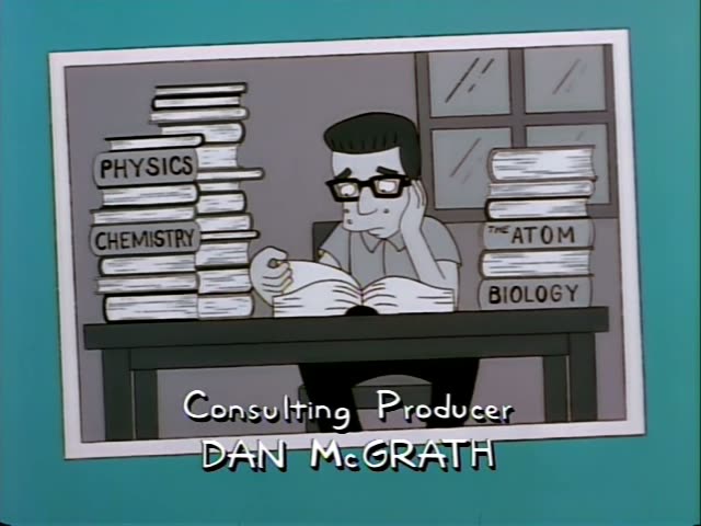 A bespectacled man with dark hair and glasses, wearing a grey shirt, sits at a desk in a drawn room, resting his cheek…