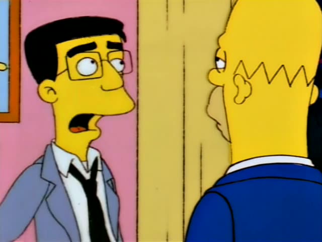 A thin man with black hair, glasses, a grey suit and a black tie stands in a doorway of a pink room, talking to a bald man…