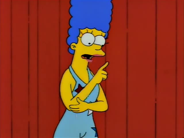 Marge Simpson stands before a red wooden backdrop, first pointing and talking, then spreading her arms wide and smiling…