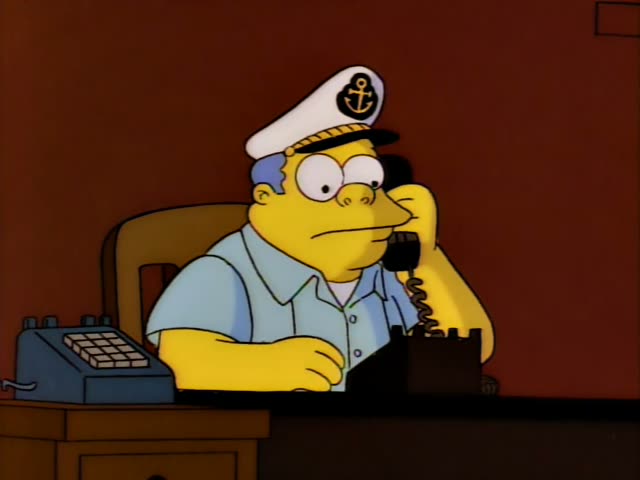 A heavy man in a white captain's cap with a gold anchor and a light blue shirt sits at a desk in a dim office…