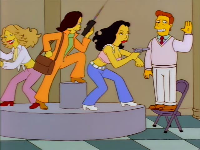 Troy McClure stands on a folding chair on a checkered floor while a dark-haired woman in a cropped top and blue jeans aims…