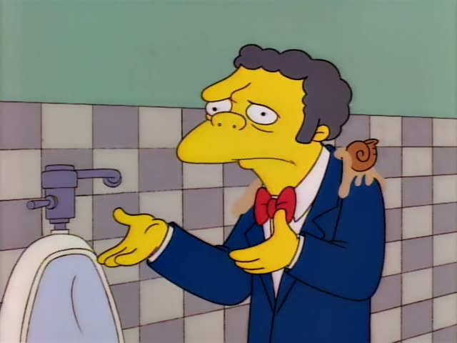 A man in a blue suit with a red bow tie stands in a tiled restroom beside a white urinal, gesturing with his hands…
