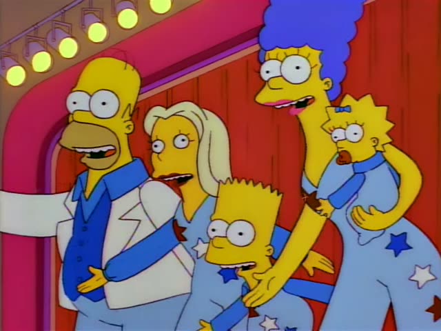 Homer in a white jacket over a blue vest, a blonde woman, Bart, and Marge holding Maggie perform on a stage before a red…