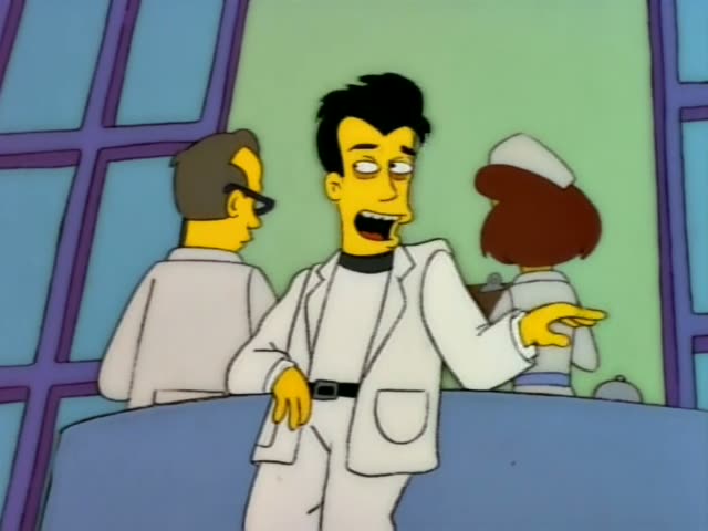 A slim man with spiky black hair, dark circles under his eyes, a white coat, white trousers and a black collar stands…
