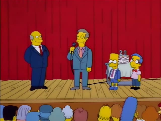 Principal Skinner stands center stage holding a microphone in front of a red curtain while a bald man in a dark suit and red…