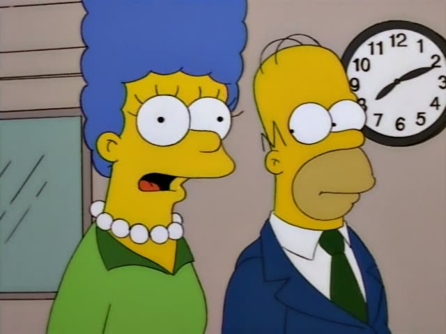 Homer stands beside Marge, who wears a green dress and a pearl necklace, in a room with a wall clock and framed certificates