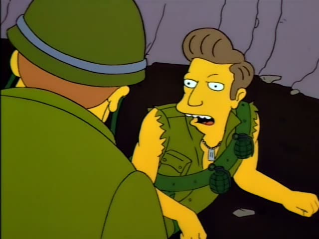 Troy McClure grenades on his sleeveless green vest and dog tags, sits on the ground and talks with a helmeted soldier…