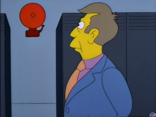 A man in a blue jacket, orange tie and purple shirt, with light brown hair, stands in a school hallway lined with grey…