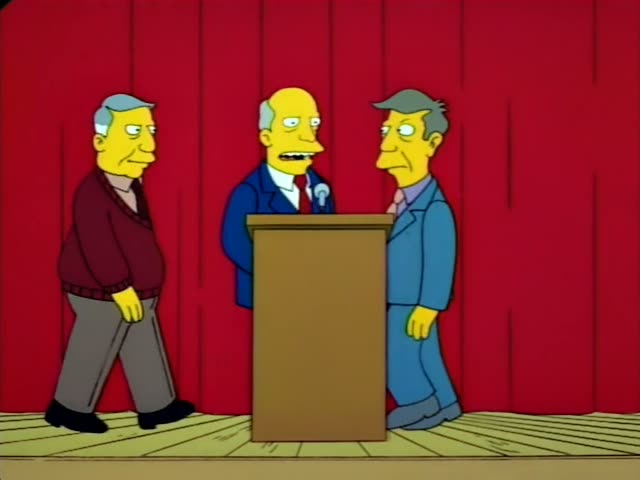 Mr. Burns walks onto a red-curtained stage toward a wooden podium, while Principal Skinner in his blue suit and an unnamed…