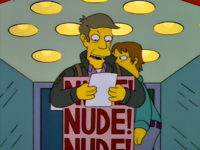 A man with short brown hair and a dark jacket, wearing a sandwich board that reads NUDE