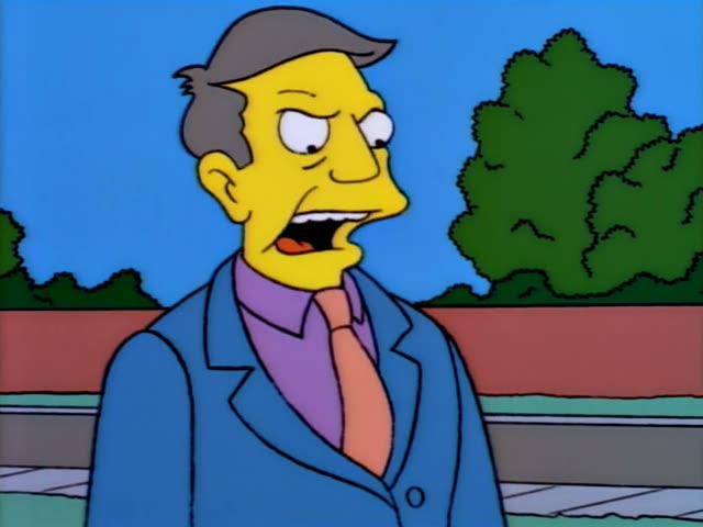 Principal Skinner in his grey-haired suit stands on a schoolyard beside a low wooden table with Bart crouched on it…