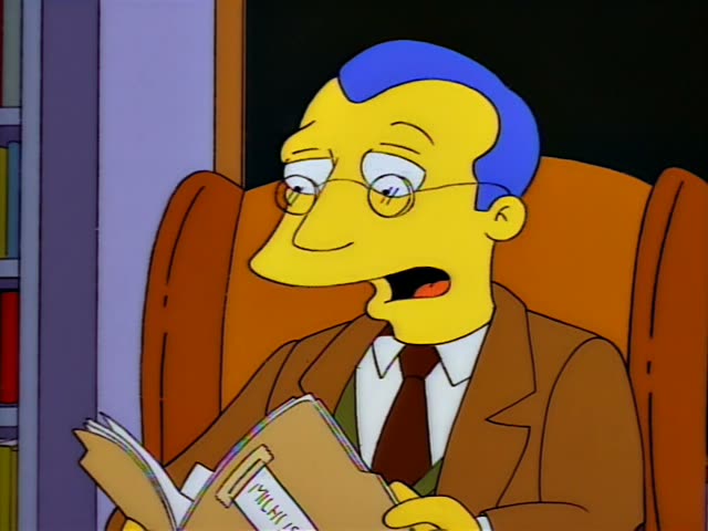 A bespectacled man with blue hair in a brown suit and green sweater sits in an orange armchair in an office, flipping…