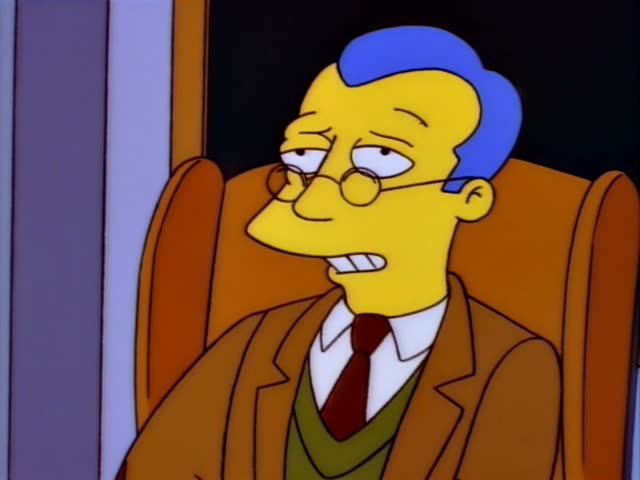 A tall-haired, thin man with blue hair, round glasses, a brown jacket, green sweater and red tie sits behind a desk…