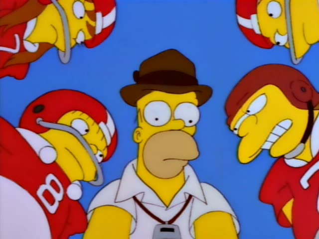 Homer Simpson huddles with a team of red-jerseyed kids in helmets on a grass field, with two onlookers seated on bleachers…