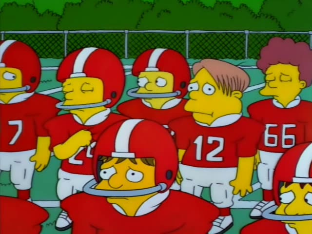 Homer Simpson stands on wooden bleachers under a blue sky and addresses a line of red-helmeted football players in red…