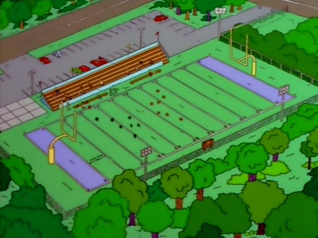 Homer stands on the sideline in front of the wooden bleachers while Ned Flanders lurks behind him and a blond boy in a red…