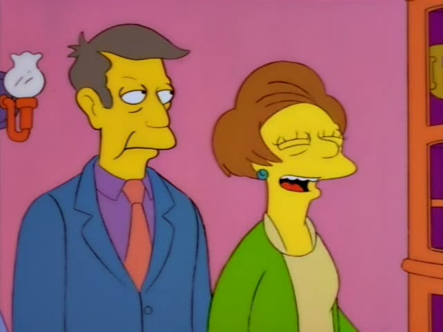 Mrs. Krabappel grins and gestures with her hands beside a neat-haired man in a blue suit and orange tie, who looks…