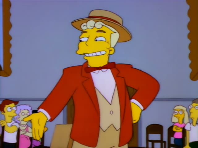 A stocky, white-haired man in a red jacket, red bow tie, tan vest and straw boater hat with a red band stands before…