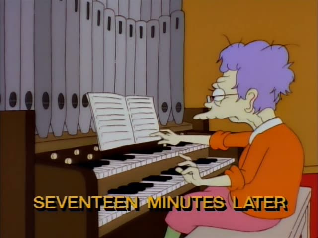A lavender-haired woman in glasses, an orange sweater and a pink skirt plays a two-manual organ, her hands moving…