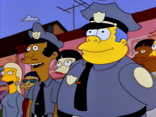 Chief Wiggum stands at the front of a row of uniformed officers and onlookers outside a pink building, smiling…