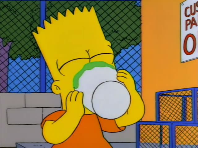 Bart stands by a chain-link fence in front of an orange wall with a partly visible sign reading "CUS