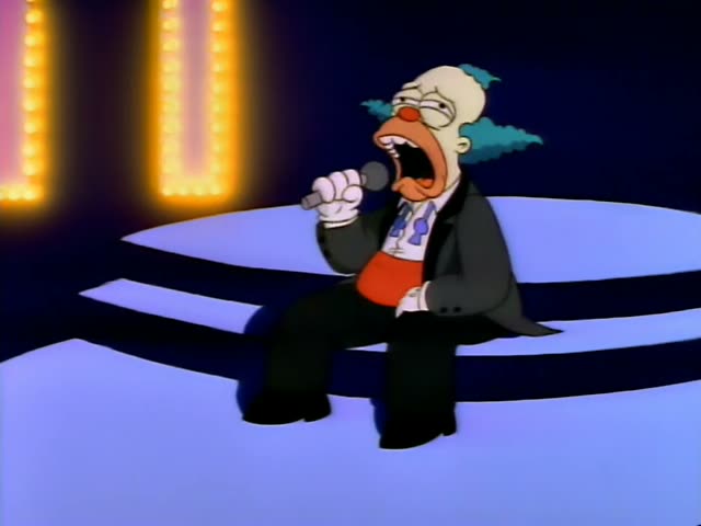 Krusty the Clown sits on a stage edge under glowing marquee lights and sings into a handheld microphone