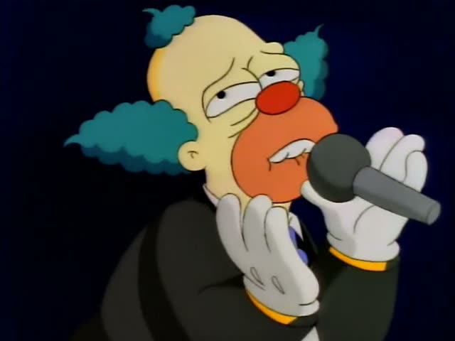 Krusty the Clown sings into a microphone while seated on the edge of a striped stage platform beneath marquee lights