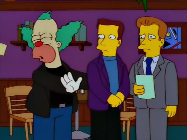 Krusty the Clown stands in a purple-walled room beside two men: a brown-haired man in a purple jacket and blue turtleneck…