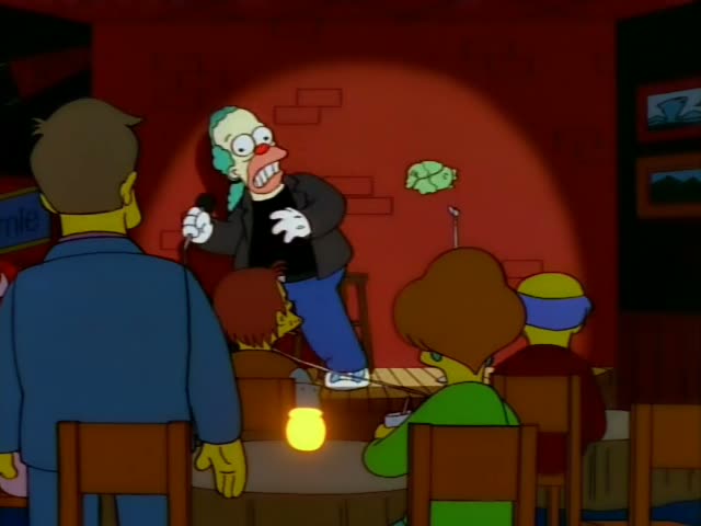 Krusty the Clown performs stand-up under a spotlight against a red brick wall in a bar, holding a microphone…