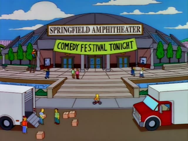 Bart Simpson first appears riding a small bike outside the Springfield Amphitheater, which has a banner…