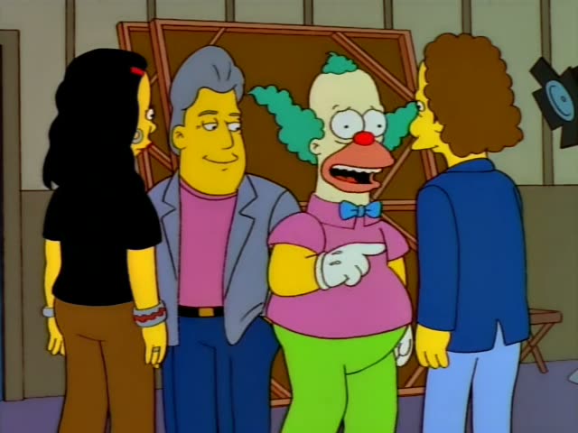 Krusty the Clown stands in a studio set with a wooden frame and a stage light, flanked by a grey-haired man in a grey jacket…