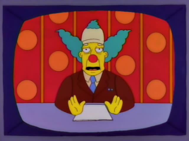 Krusty the Clown sits behind a desk in front of a red and orange polka-dot backdrop, shown on a television screen…