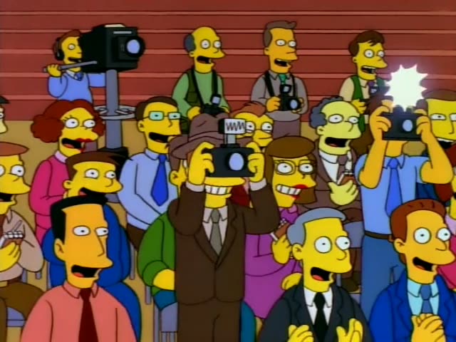 Krusty the Clown stands against an orange wooden wall clutching a handheld microphone while a crowd of reporters…