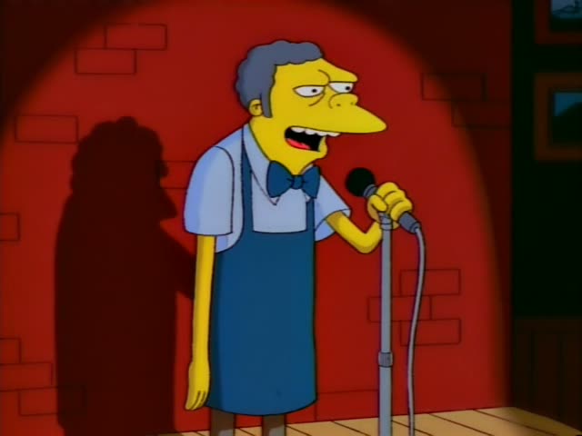 Moe stands at a microphone on a red brick-walled stage in the first several moments, gesturing and speaking with a sneer