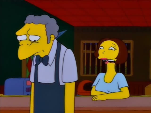 Moe stands in the bar while a brown-haired woman in a light blue top leans on the counter, gesturing with one hand…
