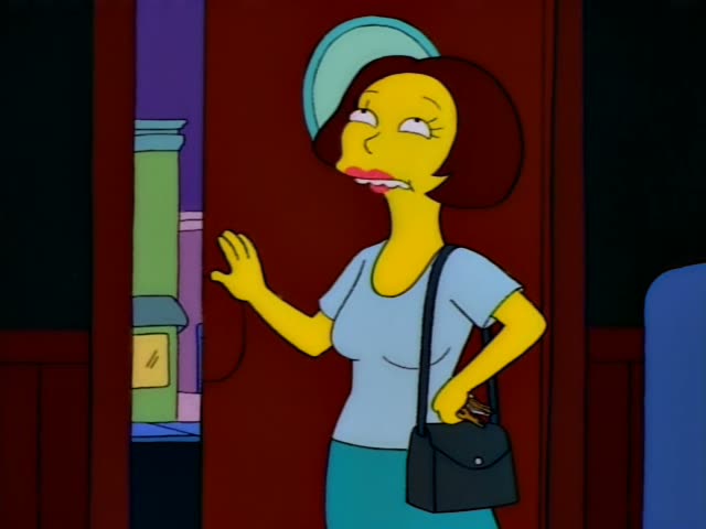 A one-off woman with a short reddish-brown bob, a light blue top, a teal skirt and a black handbag stands in a doorway…