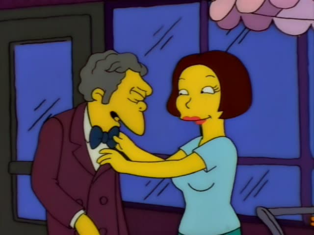 A grey-haired man in a maroon suit and blue bow tie stands outside a blue-windowed building with a pink awning while a woman…
